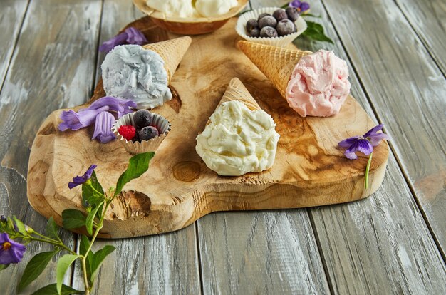 Helado de vainilla, arándano y frambuesa en conos de waffle sobre tabla de madera con frutos del bosque y flores.