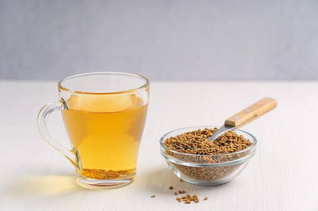 Helba oder goldener Tee aus einjähriger Bockshornkleepflanze, serviert in einer Glastasse mit einer Schüssel mit Samen und einem Löffel