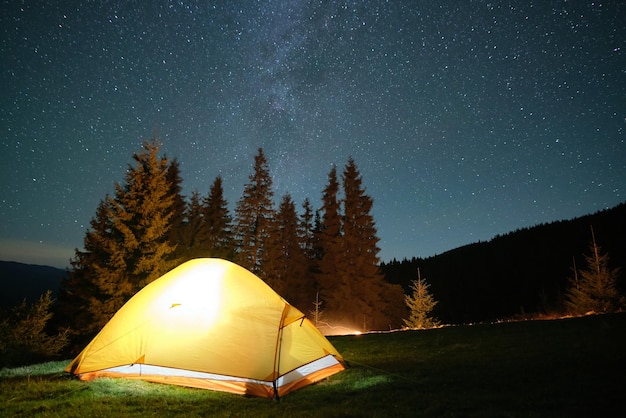 Hell beleuchtetes Touristenzelt, das auf dem Campingplatz in dunklen Bergen unter Nachthimmel mit funkelnden Sternen leuchtet Aktives Lifestyle-Konzept