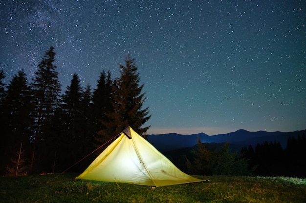 Hell erleuchtetes Campingzelt, das auf dem Campingplatz in dunklen Bergen unter Nachtsternen leuchtet Aktiver Lebensstil und Reisekonzept