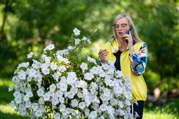 Hell gekleidete blonde reife Frau, die auf dem Smartphone im Online-Kommunikationskonzept des Gartens spricht