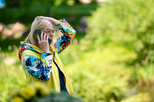 Hell gekleidete blonde reife Frau, die auf dem Smartphone im Online-Kommunikationskonzept des Gartens spricht