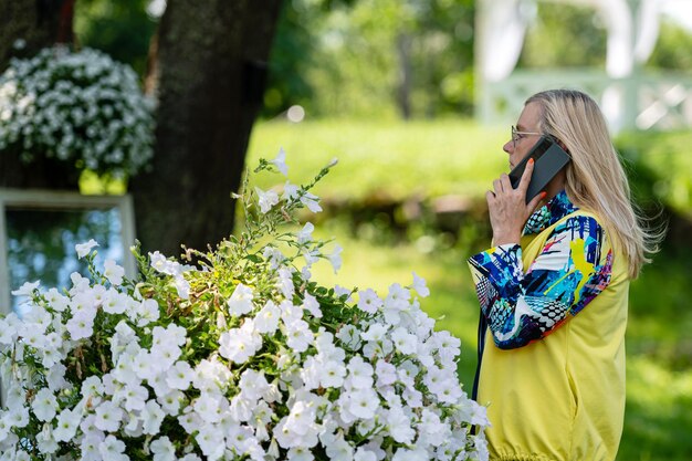 Hell gekleidete blonde reife Frau, die auf dem Smartphone im Online-Kommunikationskonzept des Gartens spricht