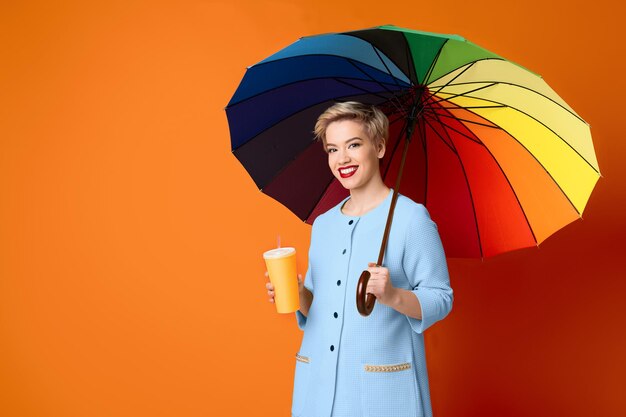 Helle blonde Frau mit Kaffeepapiertasse unter Regenbogenregenschirm auf orangem Hintergrund, Kopienraum