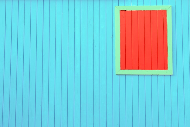 Helle, farbenfrohe blaue Holzwand des Hauses mit geschlossenem orangefarbenem Fenster in grünem Rahmen als strukturierter abstrakter Hintergrund, Kopierraum