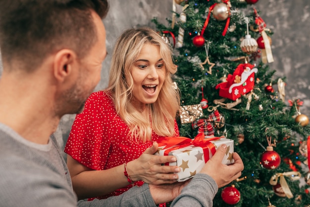 Helle, glückliche Emotionen der jungen Frau erhalten ein Geschenk vom Mann vor dem Hintergrund des in rot-grünen Tönen dekorierten Weihnachtsbaums. Lifestyle-Foto