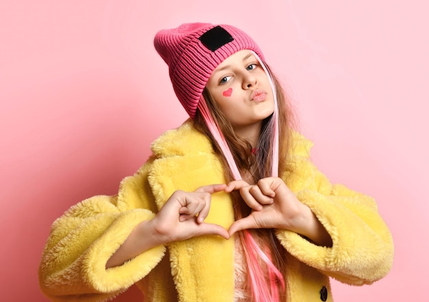 Helles jugendlich Mädchen, das Herz zur Kamera zeigt, während es auf rosa Hintergrund steht Mädchen in einem gelben Pelzmantel mit Hut und mit einem gemalten Herzen auf ihrem Gesicht zeigt ein Zeichen der Liebe und sendet Luftküsse
