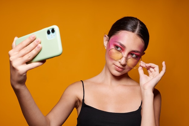 Helles Make-up der jungen schönen Frau, das schwarzes Trikot mit einem Telefon in der Hand aufwirft, gelber Hintergrund unverändert