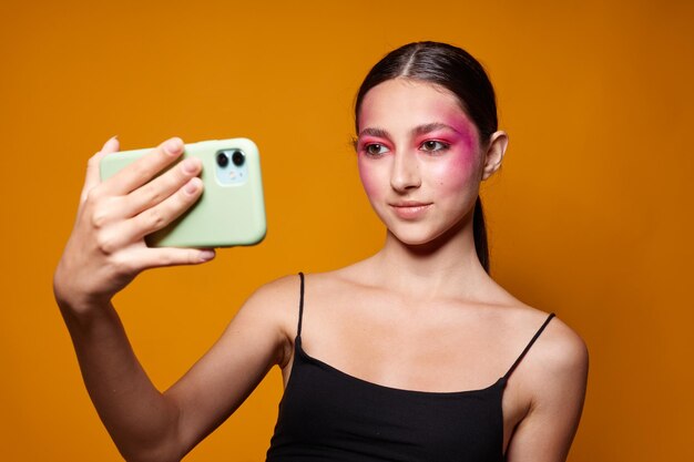 Helles Make-up der sexy Brunettefrau, das schwarzes Trikot mit einem Telefon in der Hand gelben Hintergrund unverändert aufwirft