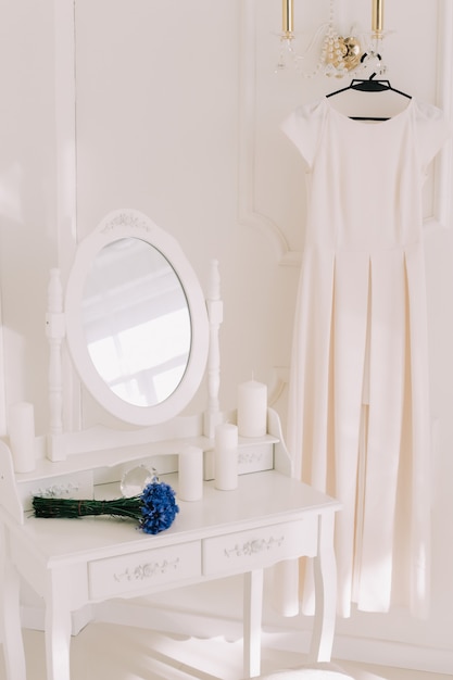 Helles romantisches Interieur mit einem Hochzeitskleid auf einem Kleiderbügel, einem Blumenstrauß und einem Schminktisch mit einem Spiegel