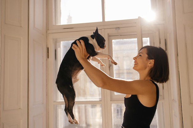 Hellhäutiges junges brünettes Mädchen hält an einem sonnigen Tag am Fenster einen schwarzen und weißen Hund vor sich Liebe zwischen Hund und Besitzer Konzept