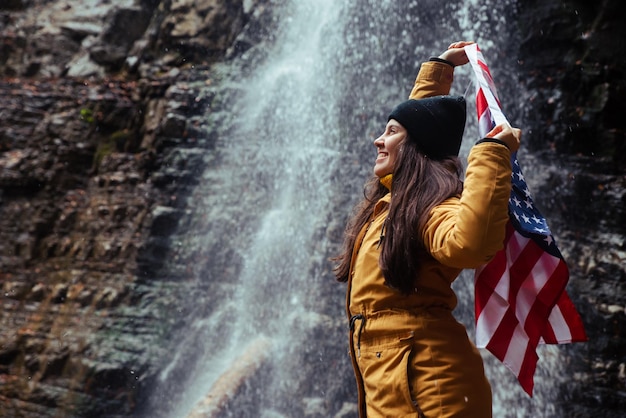 Hembra adulta caucásica joven con bandera de estados unidos frente a cascada