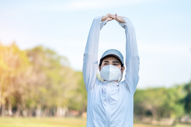 Hembra adulta joven vistiendo N95 mascarilla protectora durante el estiramiento muscular en el parque al aire libre