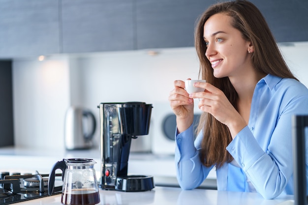 Hembra atractiva feliz que bebe el café aromático fresco caliente después de preparar el café usando la cafetera en la cocina en casa. Batidora de café y electrodomésticos para hacer bebidas calientes