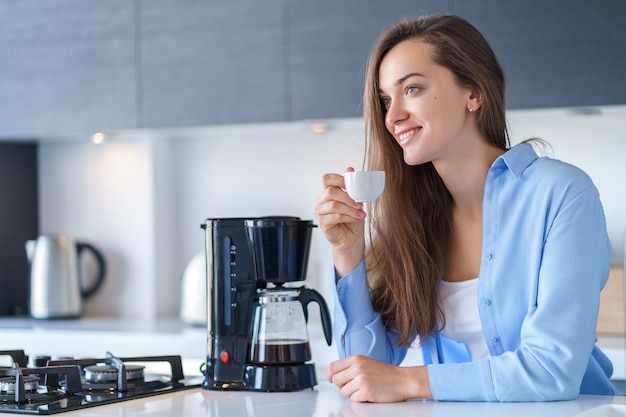 Hembra atractiva feliz que bebe el café aromático fresco caliente después de preparar el café usando la cafetera en la cocina en casa. Batidora de café y electrodomésticos para hacer bebidas calientes