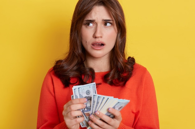 Hembra europea adorable desconcertada con suéter naranja que se encuentran aisladas sobre una pared amarilla con la boca abierta y mirando a otro lado, sosteniendo el dinero en efectivo en las manos y pensando cómo gastar.