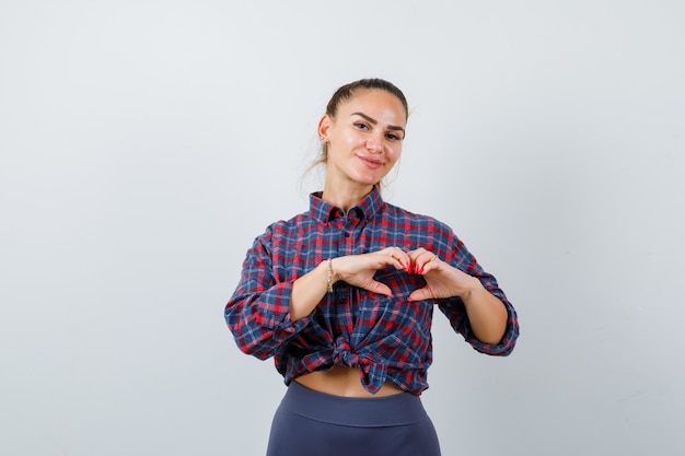 Hembra joven en camisa a cuadros, pantalones mostrando gesto de corazón y luciendo lindo, vista frontal.