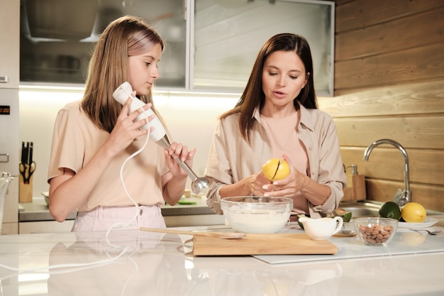 Hembra joven cortando limón en dos mitades sobre un tazón con leche mientras su hija va a mezclar los ingredientes con una batidora eléctrica