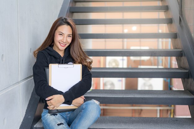 La hembra joven del estudiante se sienta en las escaleras con la carpeta y el libro de copia en área del campus