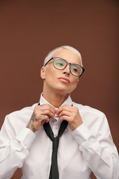 Hembra joven con pelo rubio teñido corto, piercing en la nariz y tatuajes haciendo botones en el cuello de la camisa blanca