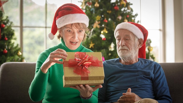 Hembra madura en el presente de apertura del sombrero de Santa mientras está sentado en el sofá cerca del marido senior feliz durante la celebración de Navidad en casa