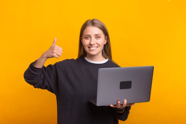 La hembra sonriente está mostrando el pulgar hacia arriba mientras sostiene su computadora portátil.