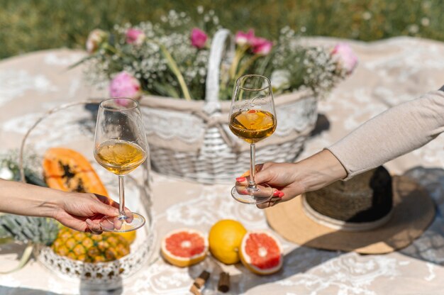 Hembras irreconocibles extendiendo los brazos para mostrar dos copas con vino blanco. Picnic de manta en ambiente de fondo con frutas y flores tropicales.