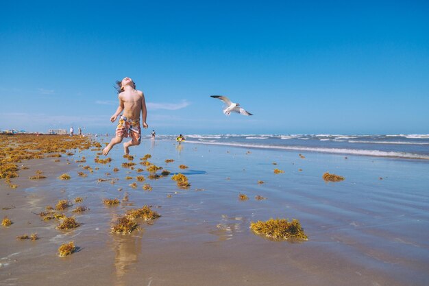 Hemdloser Junge springt auf dem Strand gegen den blauen Himmel