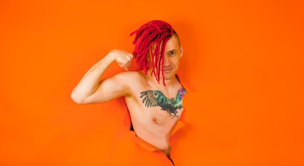Hemdloser Mann mit roten Dreadlocks und mit Vogeltattoo auf der Brust, der Bizeps zeigt und durch ein Loch in einer orangefarbenen Wand im Studio in die Kamera blickt