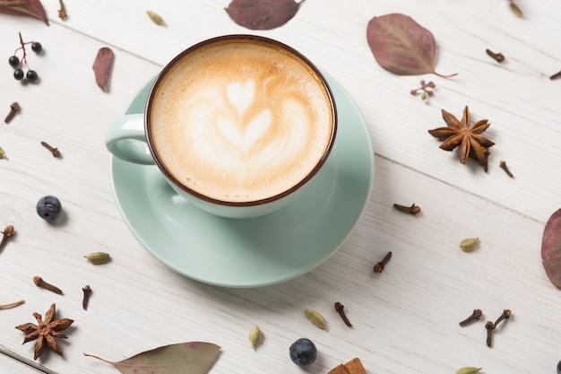 Herbst Cappuccino Zusammensetzung. Blaue Kaffeetasse mit Schaum, Nelken, Schlehe, getrockneten Blättern am weißen Holztisch. Herbst heiße Getränke, Café und Bar Konzept