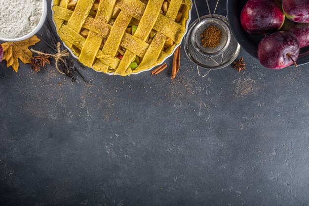 Herbstapfelkuchen, der Hintergrund kocht. Backhintergrund mit rohem traditionellem klassischem Apfelkuchen, Backzutaten und Utensilien, schwarzer Betonhintergrundkopierraum