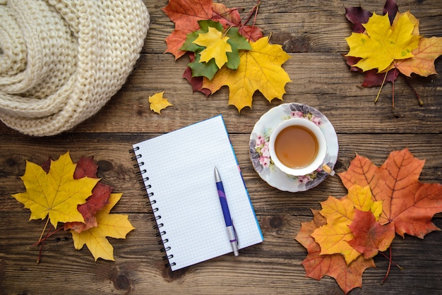 Herbstbild von Gelbblättern, von Tasse Tee, von Schal und von Blatt Papier mit Stift auf hölzernem Hintergrund