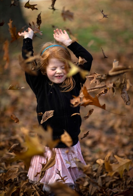Herbstblätter fallen auf ein Mädchen mit erhobenen Armen, das im Park steht.