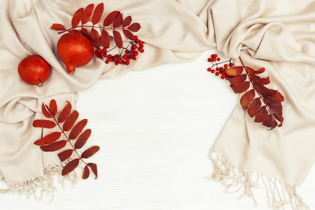 Herbsthintergrund mit roter Farbe der Ebereschenblätter und Ebereschenbeeren auf weichem herbstlichem Frauenkleidungsschal, orange Pampkins auf weißem Holz mit Kopienraum. Draufsicht. Flach liegen