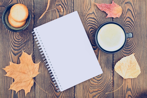 Herbstkomposition mit einem leeren Tagebuch mit trockenen Herbstblättern im Frühling eine Tasse Kaffee und Kekse auf einem Holzhintergrund Flacher Kopienraum