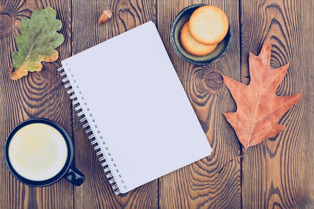 Herbstkomposition mit einem leeren Tagebuch mit trockenen Herbstblättern im Frühling, einer Tasse Kaffee und Keksen an