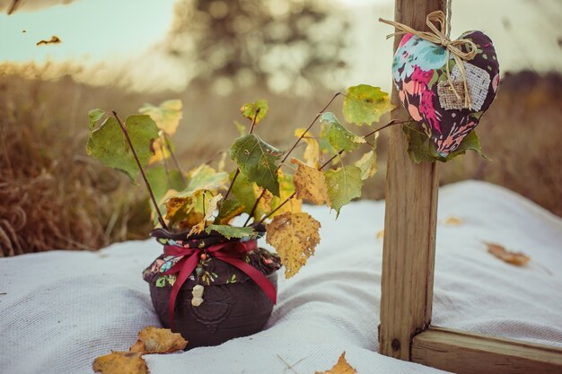 Herbstkomposition mit Holzrahmen, weichen Herzkissen und einer Vase mit gelben Blättern auf einer Decke draußen. Herbstfotobereich und handgemachte Dekoration