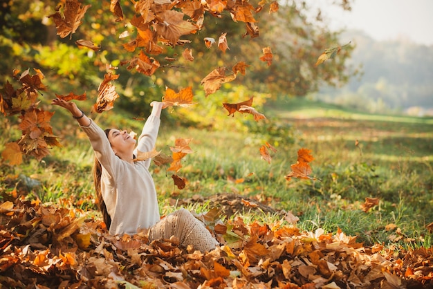 Herbstlaub fällt auf glückliche junge Frau im Wald Die Farben und die Stimmung des Herbstes Schöne junge Frau, die Blätter in einem Park wirft Herbstkonzept Glückliches lächelndes Mädchen mit natürlichen roten Haaren