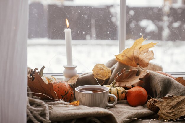 Herbstliches Stillleben. Warme Tasse Tee, Kürbisse, Herbstblätter, Kerzenlichter auf einem bequemen braunen Schal auf der Fensterbank. Hygge-Herbst-Wohnkultur. Fröhliches Thanksgiving