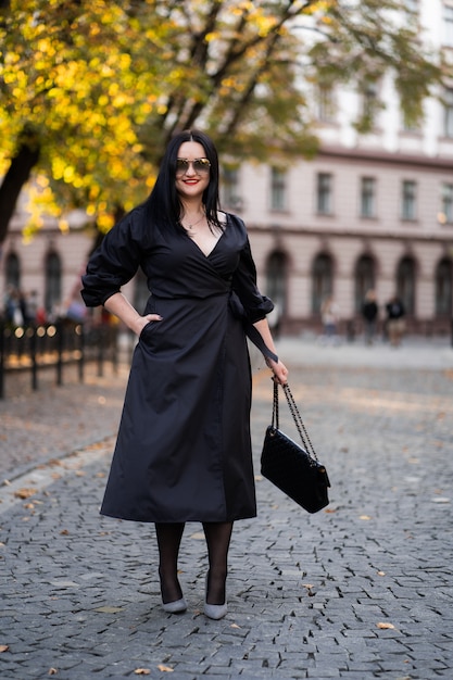 Herbstmode. Das Mädchen mit den roten Lippen im modischen stilvollen schwarzen Kleid und in der Sonnenbrille, herbstlicher Lebensstil auf dem Hintergrund der verschwommenen gelbgrünen Bäume im Park.