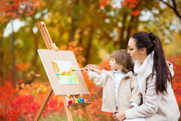 Herbstnatur, Mutter und Tochter malen ein Bild in einem Park aus Herbstblättern, malen ein kleines Kind, Kinderkreativität.