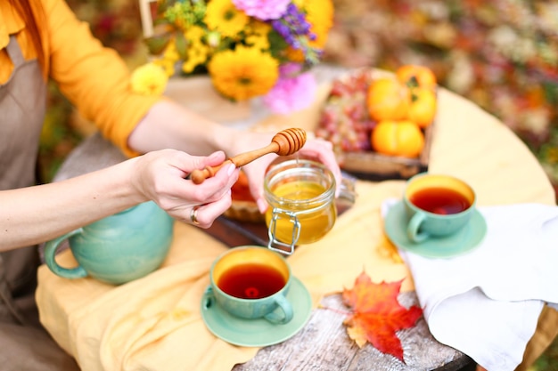 Herbstpicknick Frau im gelben Kleid und Leinenschürze trinkt Tee aus der Tasse am Holztisch im Garten Schöner Wasserkocher Tischdecke Honig mit Löffel Apfelkuchenernte Kaki Trauben Ahornblatt