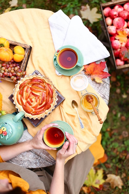 Herbstpicknick Frau im gelben Kleid und Leinenschürze trinkt Tee aus der Tasse am Holztisch im Garten Schöner Wasserkocher Tischdecke Honig mit Löffel Apfelkuchenernte Kaki Trauben Ahornblatt