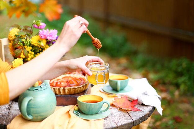 Herbstpicknick Teeparty mit schönen Kesselbechern am Holztisch im Garten Erntefest Honig mit Stocklöffel Apfelkuchen Kaki Trauben Ahornblatt Blumen gelbe Leinentischdecke