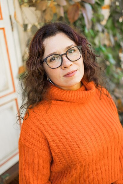 Herbstporträt einer attraktiven jungen Frau mit stylischer Brille in einem gestrickten, modischen orangefarbenen Pullover auf dem Hintergrund von Blättern Mädchen geht in der Stadt spazieren