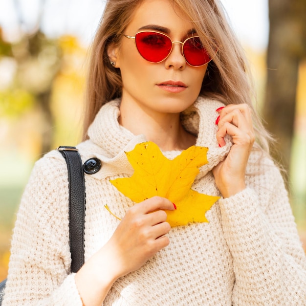 Herbstporträt einer jungen schönen Frau mit roter Mode-Sonnenbrille im weißen Pullover hält ein farbiges gelbes Herbstblatt und geht im Park spazieren