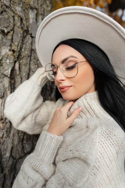 Herbstporträt einer schönen, stilvollen Frau mit Vintage-Brillenmütze und Strickpullover in einem Herbstpark in der Nähe eines Baums