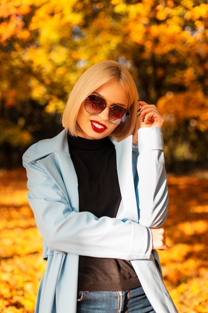 Herbstporträt eines schönen jungen blonden Mädchens mit Sonnenbrille in einem hellblauen Mantel mit schwarzem Pullover geht in einem Herbstpark mit goldenem Herbstlaub