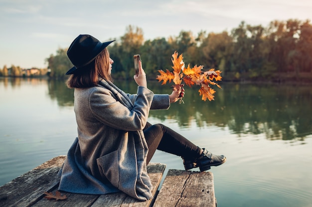 Herbstsaison. Frau, die Fotos von Niederlassungen mit Blättern unter Verwendung des Smartphone auf Flusspier macht