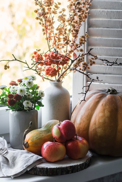 Herbststillleben am Fenster ein Kürbis eine Vase mit trockenen Zweigen und Eberesche reife saftige Äpfel Herbstkonzept eines gemütlichen Interieurs